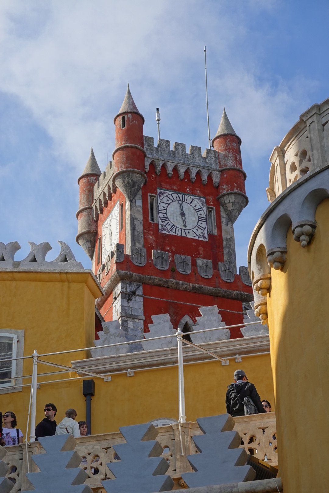 Red and grey battlemented clock tower of Pena Palace framed by yellow walls against a cloudy sky