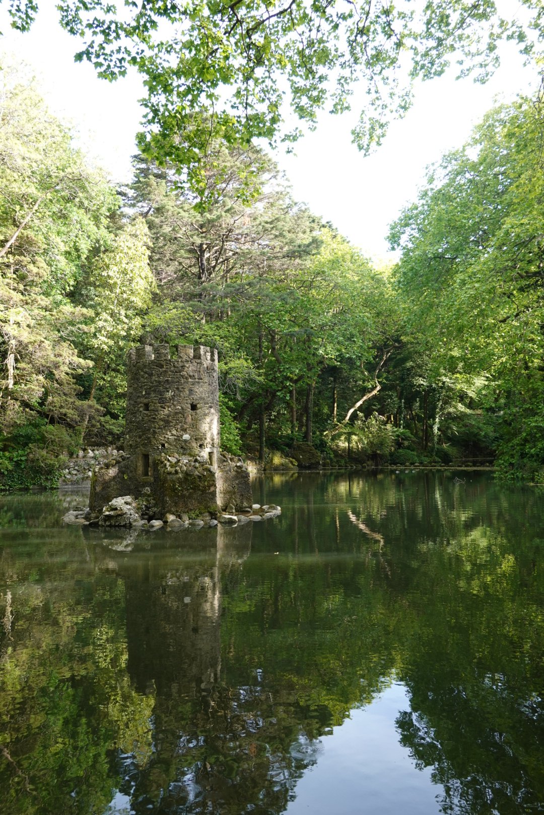 A ruined stone tower on a small island reflected in the still green lake of Quinta da Regaleira