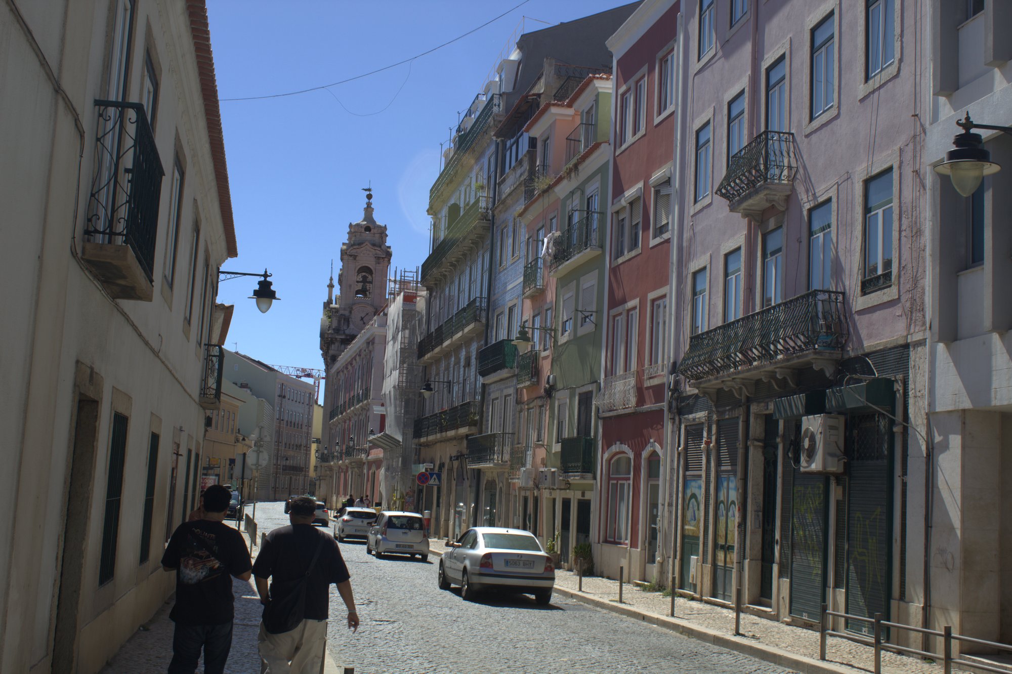 Two people walking down a cobblestone Lisbon street lined with pastel-coloured buildings and wrought-iron balconies