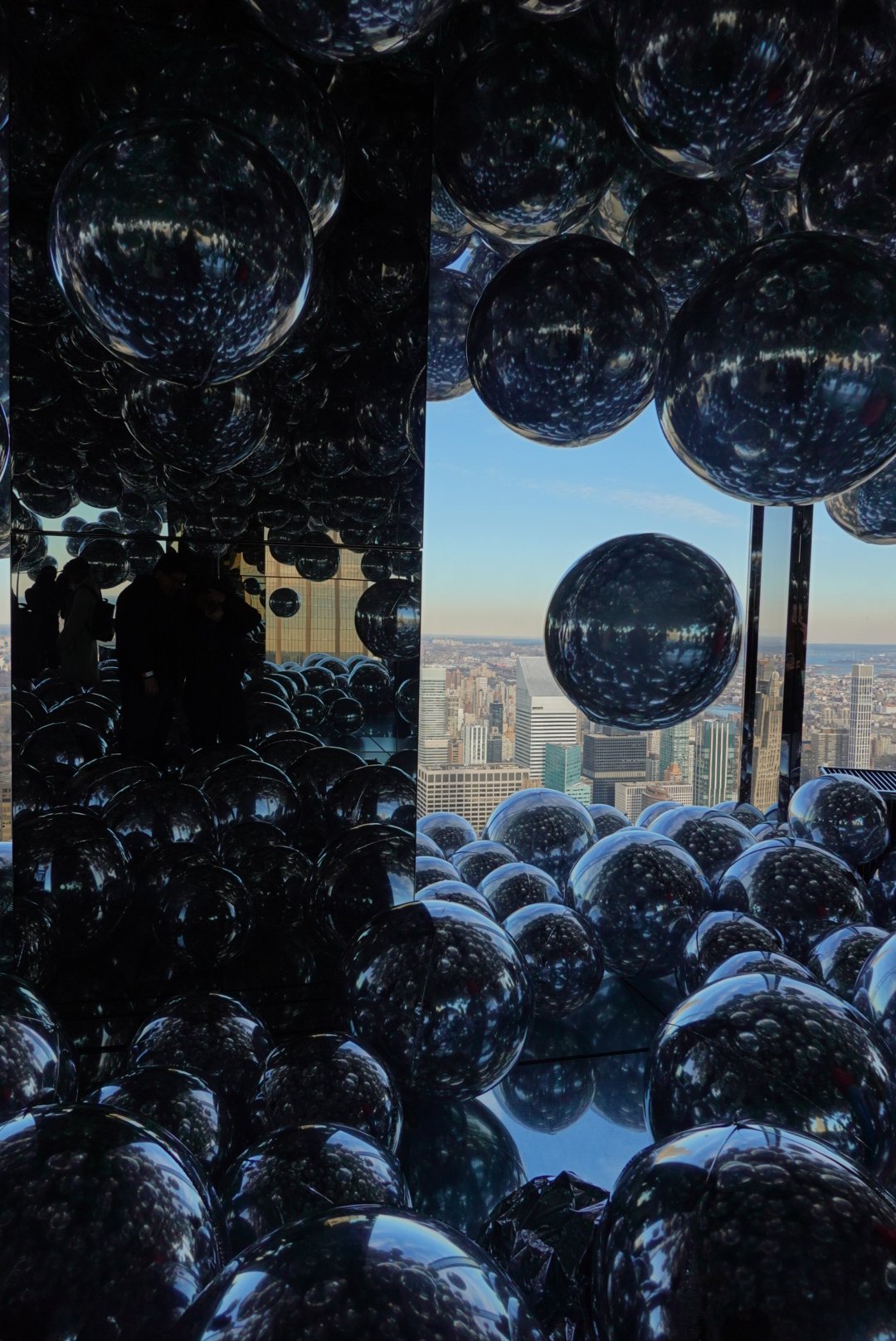 Mirrored spheres filling a room at an observation deck, Manhattan skyline visible through the window